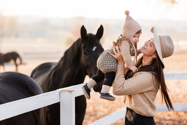 De gelukkige jonge moeder speelt met weinig babydochter dichtbij de paarden in de herfstpark