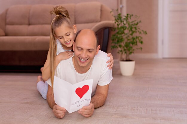 De gelukkige jonge papa houdt een kaart van kleine vrolijke dochters op vaderdag tijdens huisvakantie