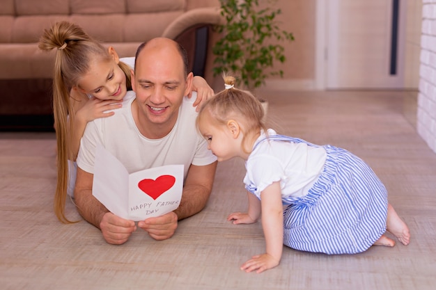 De gelukkige jonge papa houdt een kaart van kleine vrolijke dochters op vaderdag tijdens huisvakantie