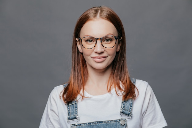 De gelukkige jonge vrouw met blije gelaatsuitdrukking, heeft een gezonde huid, draagt optische glazen