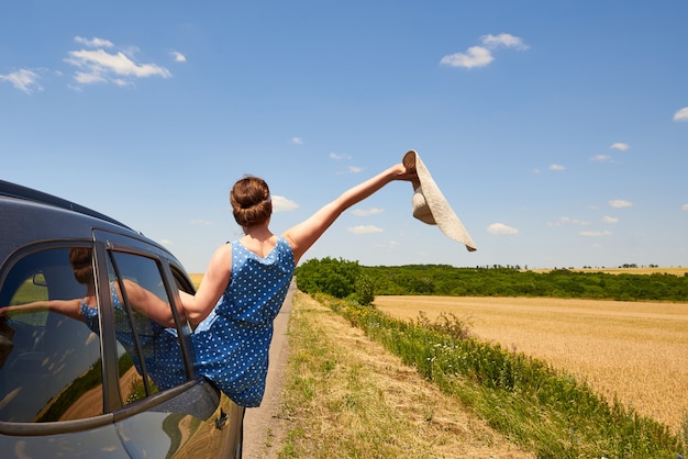 De gelukkige jonge vrouw met een hoed kijkt uit het autoraam op de weg.