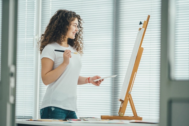 De gelukkige krullende vrouw schildert een schilderij op de ezel