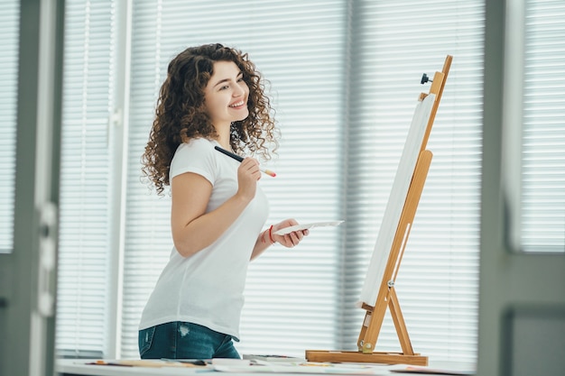 De gelukkige krullende vrouw schildert een schilderij op de ezel