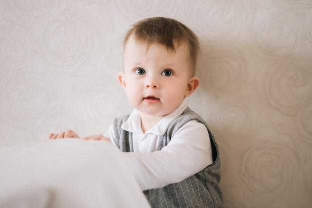 De gelukkige leuke jongen in het kostuum van een heer bevindt zich dichtbij de steun op een lichte achtergrond, proberend op te staan, lerend te lopen.