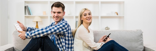 De gelukkige man en vrouw met een telefoon en een tablet zitten op de bank