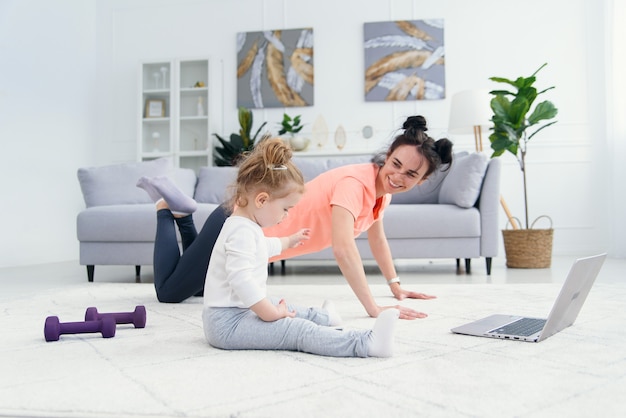 De gelukkige moeder die ochtendoefeningen in yoga doen stelt terwijl haar weinig dochter die thuis spelen. Jonge schattige moeder plezier beoefenen van meditatie ontspannen op stressvrij weekend met babymeisje.