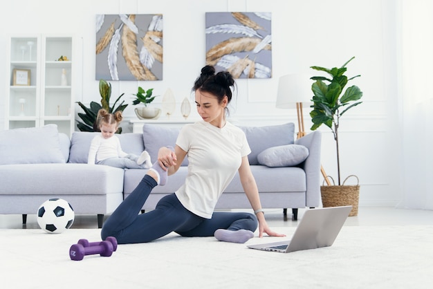 De gelukkige moeder die ochtendoefeningen in yoga doen stelt terwijl haar weinig dochter die thuis spelen. jonge schattige moeder plezier beoefenen van meditatie ontspannen op stressvrij weekend met babymeisje.