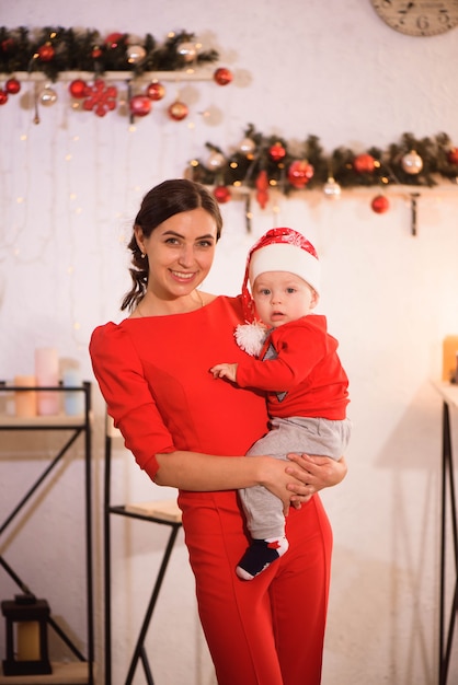 De gelukkige moeder en de aanbiddelijke baby in Kerstmanhoed vieren Kerstmis. Nieuwjaars vakantie. Peuter met moeder in de feestelijk ingerichte kamer