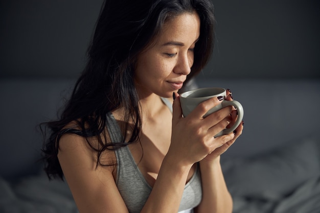 De gelukkige mooie aziatische vrouw houdt een kop en geniet van haar ochtendkoffie