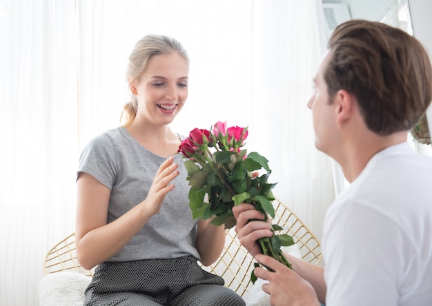 De gelukkige mooie jonge kaukasische vrouw kreeg bosrozen met verrassing van de mens terwijl thuis op bank in slaapkamer zit.