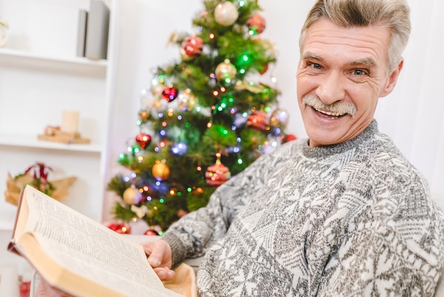 De gelukkige oude man houdt een boek bij de kerstboom