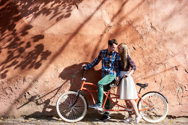 De gelukkige romantische gebaarde man en het blonde vrouw die van het toeristenpaar zich op dubbele fiets achter elkaar verenigen op achtergrond van hoge rode oude geknapperde gepleisterde muur.