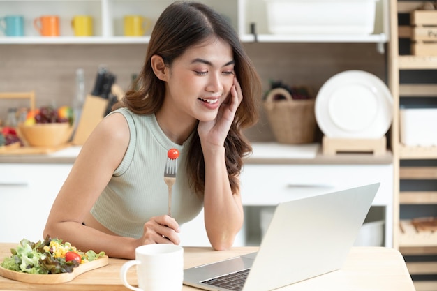 De gelukkige vrouw die van Azië salade eet met ziet computernotitieboekje in keukenruimte