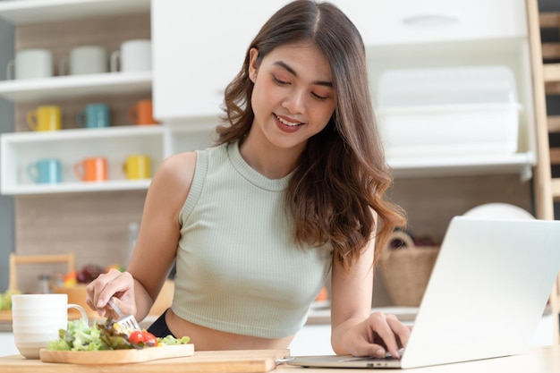 De gelukkige vrouw die van Azië salade eet met ziet computernotitieboekje in keukenruimte