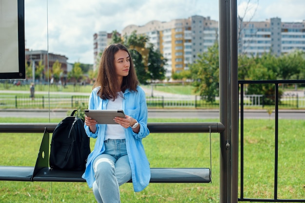 De gelukkige vrouw gebruikt een tablet of ebook op een trampost terwijl het wachten op openbaar vervoer.