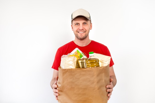 De gelukkige werknemer van de leveringsmens in schenkingen van de de handschoengreep van glb de lege rode t-shirt eenvormige houdt een document zak met voedsel op wit