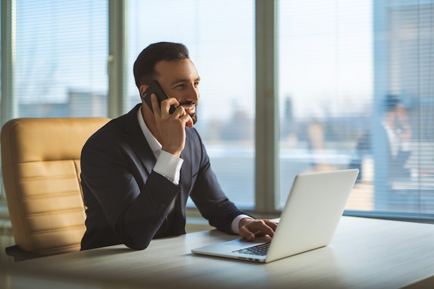 De gelukkige zakenman telefoneert aan tafel met een laptop