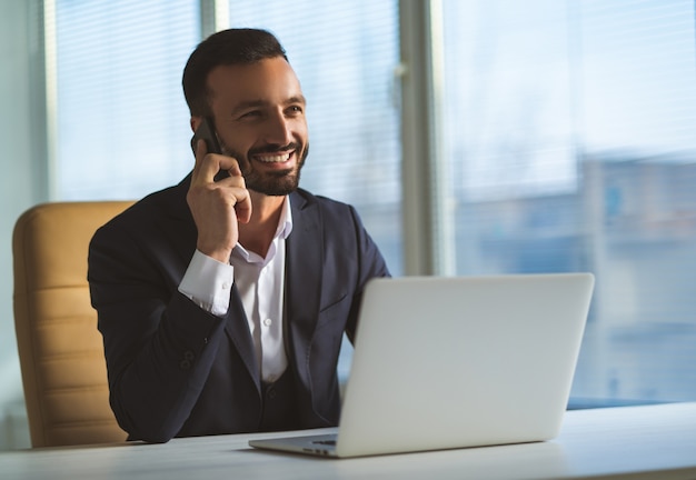 De gelukkige zakenman telefoons in de buurt van de laptop op het bureau