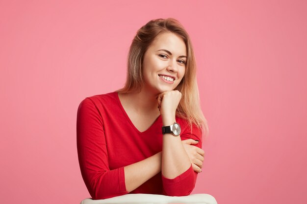 De gelukkige zekere blonde aantrekkelijke vrouw houdt hand onder kin, heeft glanzende glimlach, draagt rode sweater, die over roze muur wordt geïsoleerd. Vrij mooie vrouw poseert alleen in de studio, in een goed humeur