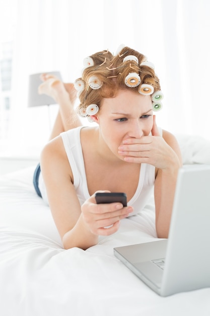 De geschokte vrouw in haarkrulspelden met cellphone bekijkt laptop in bed