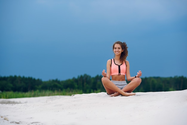 De gezonde vrouw het praktizeren yoga en mediteert op het strand in het zand dichtbij van blauwe cloudly hemel