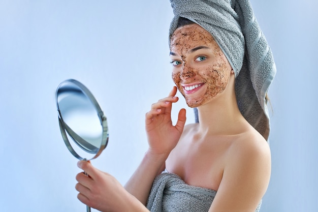 De glimlachende gelukkige vrouw in badhanddoek met natuurlijke reinigende gezichtskoffie schrobt masker na douche