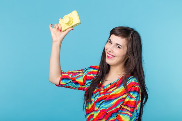 De glimlachende jonge vrouw houdt in haar handen een geel uitstekend cameramodel poseren op een blauwe muur.