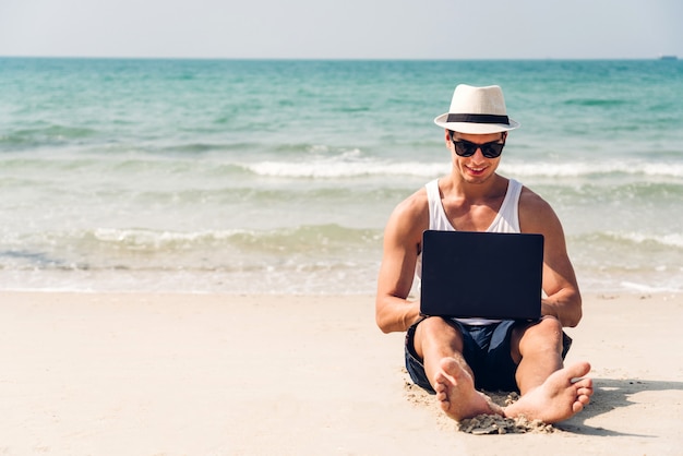 De glimlachende knappe mens ontspant in zonnebril en strohoed die aan laptop op strand werkt