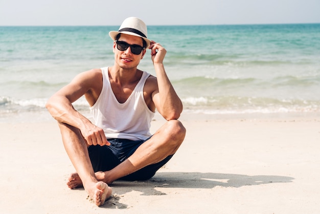 De glimlachende knappe mens ontspant in zonnebril en strohoed op het tropische strand. De zomervakanties