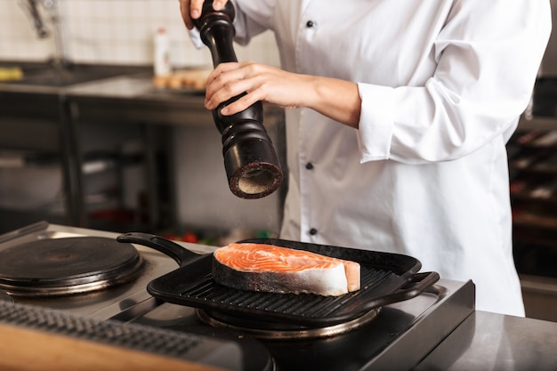 De glimlachende kok die van de vrouwenchef-kok uniform het koken van heerlijke zalmlapje vlees draagt dat zich bij de keuken bevindt, peperend