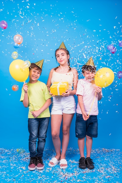 De glimlachende vrienden vieren verjaardagspartij met gift; ballonnen; en confetti over blauwe achtergrond