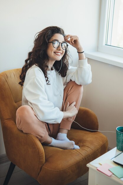 De glimlachende vrouw met krullend haar luistert naar online lessen op de laptop terwijl ze een kopje thee drinkt