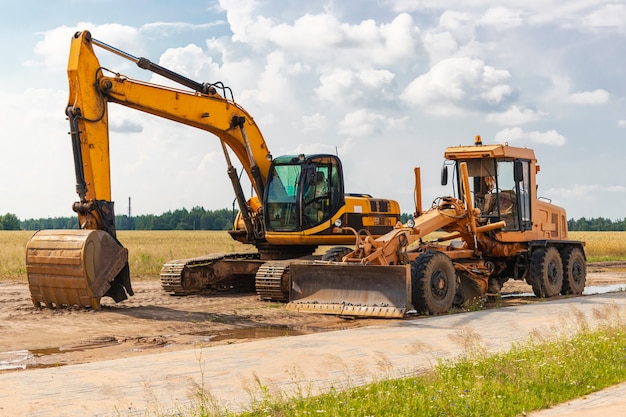 De graafmachine en de grader staan zij aan zij tegen de blauwe lucht. Grondverzetmachines voor zware constructies. Aanleg van wegen en ondergrondse communicatie. Bouwindustrie.