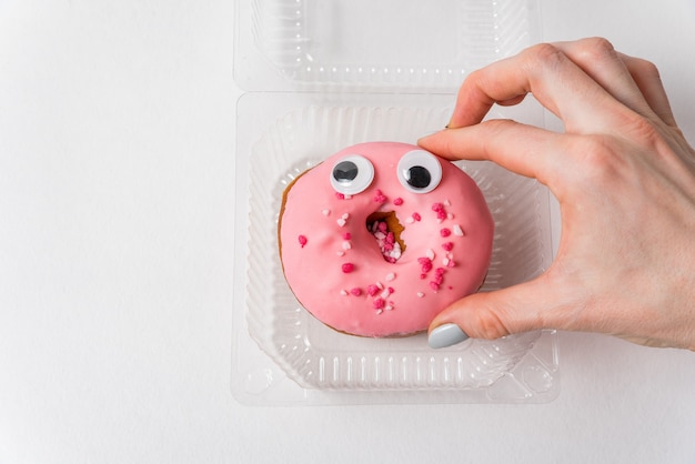De grappige roze doughnut van het schokgezicht op witte achtergrond. Vrouwelijke hand die doughnut met ogen neemt