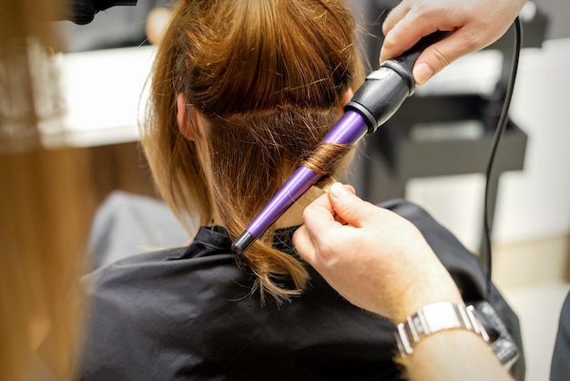 De haarstylist maakt krullenkapsel van lang bruin haar met de krultang in de kapsalon close-up