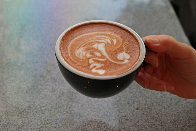 De hand die van de vrouw een kop van cappuccinokoffie met onscherpe steenlijst houden op de achtergrond