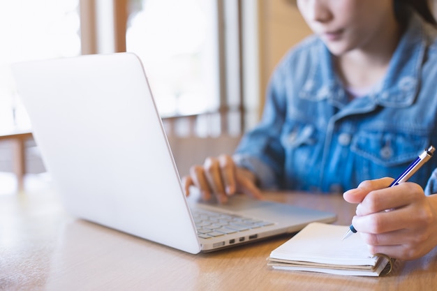 De hand die van de vrouw laptop met behulp van en in blocnote schrijft.