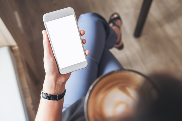 Foto de hand die van een vrouw witte mobiele telefoon met het lege desktopscherm houden terwijl het drinken van koffie