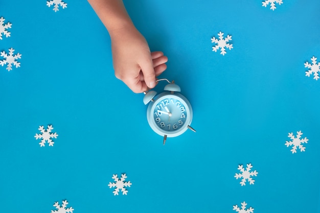 De hand die van het kind blauwe wekker houdt die vijf tot twaalf met document sneeuwvlokken toont