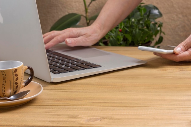 De hand typt op een tweedehands laptop en houdt een smartphonepen en een kopje koffie vast op een lichte houten tafel. Werk vanuit huis