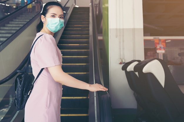 De hand van de Aziatische vrouw houdt de leuning van de roltrap vast, hoest in haar elleboog en houdt sociale afstand van andere mensen met chirurgische gezichtsmaskerbescherming - ze pendelt in het winkelcentrum