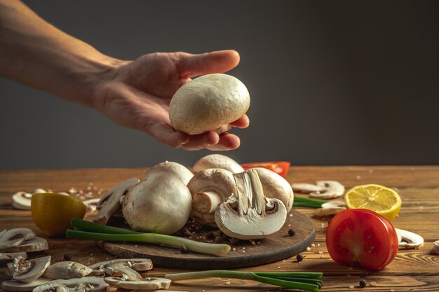 De hand van de chef houdt een verse paddenstoel boven een houten tafel met groenten en andere ingrediënten om het gerecht te koken