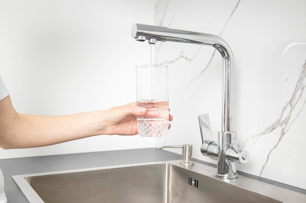 De hand van de vrouw die een glas vasthoudt, giet water uit een kraan in de keuken