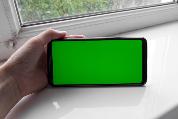 De hand van de vrouw houdt de smartphone in een horizontale positie met een groen scherm op een raam. Chroma sleutel. Bespotten.