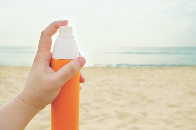 De hand van de vrouw houdt een zonnebrandspray vast in een oranje fles tegen de achtergrond van een zandstrand en zee