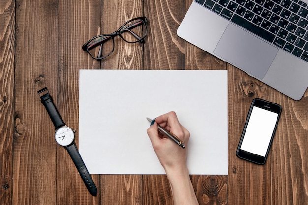 De hand van de vrouw schrijft een pen op een vel wit papier. Houten bureau tafel met laptop, telefoon, duidelijk Witboek en kantoorbenodigdheden achtergrond