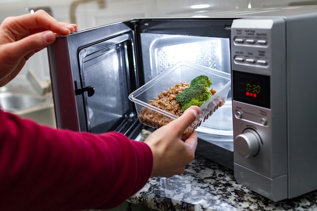 De hand van de vrouw zet plastic container met broccoli en boekweit in de magnetron