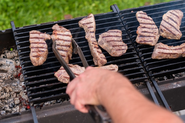 De hand van een man draait biefstuk met een tang om op een houtskoolgrill in de tuin