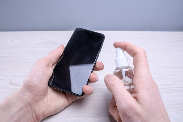 De hand van een man houdt een desinfecterende spray vast en klikt erop en desinfecteert de telefoon om verschillende oppervlakken te desinfecteren