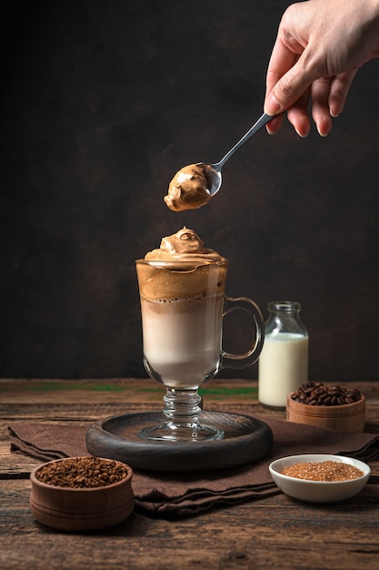 De hand van een vrouw doet koffieschuim in een glas dalgona-koffie op een bruine muur. Verticale weergave.